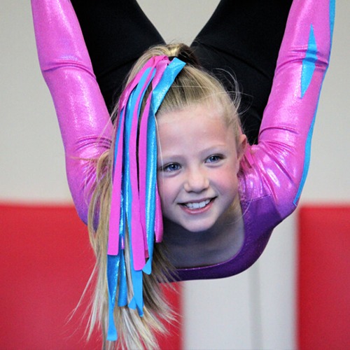 A young child hanging upside down smiling. The girl is wearing a purple, black and blue gym outfit. She has the same colours as ribbons in her hair.