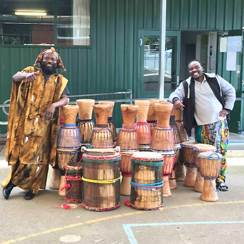 Super Mande Percussion: African Drumming Academy. Two teachers are standing next to several African drums smiling at the camera.