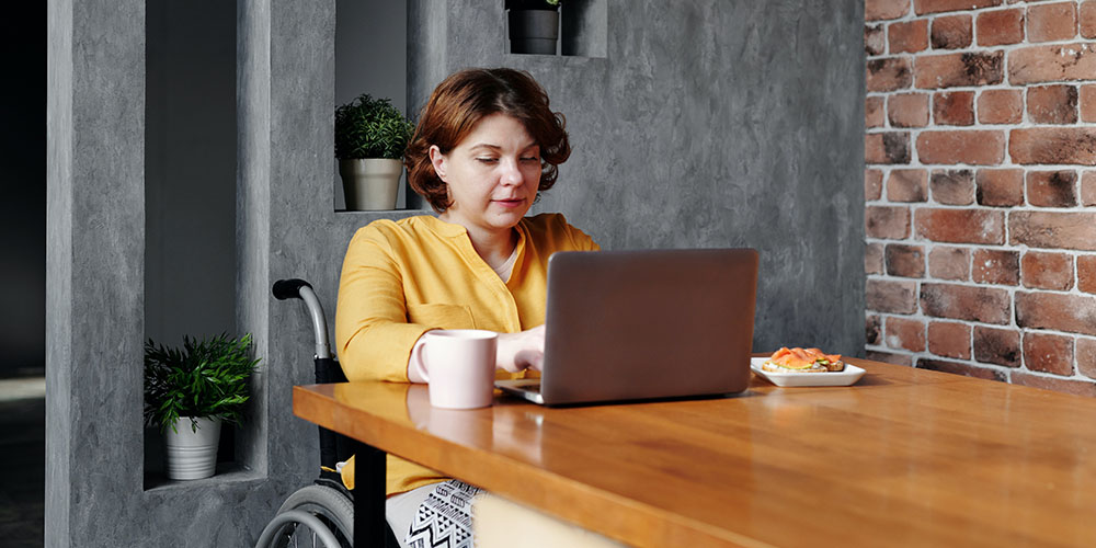 A woman with reddish hair and wearing a yellow top has breakfast at a long wooden table while working on her laptop. She is sitting in a wheelchair.