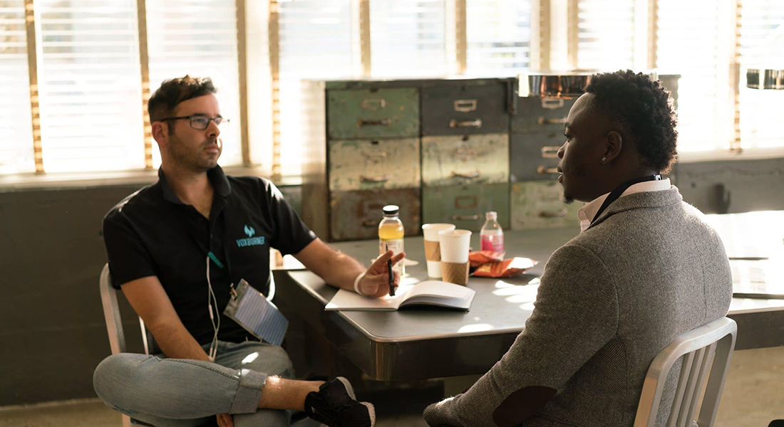 A white man with glasses and a man of African appearance in a grey jacket sit at a table talking. There are coffee cups and drinks in plastic bottles on the table.