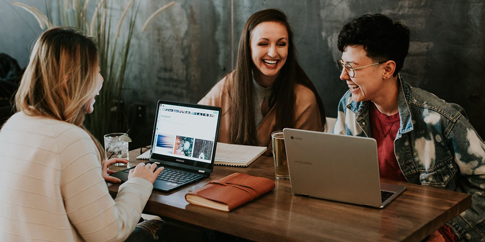 Three women sit around a wooden table laughing with their laptops open in front of them.