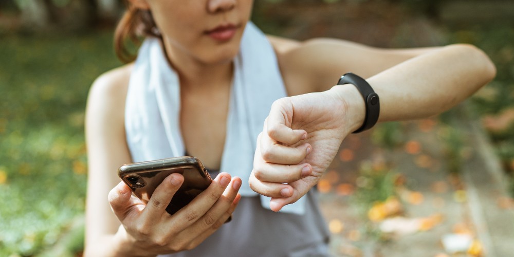 A woman hold her phone in her hand while checking her watch after a workout. She has a gym towel draped over her shoulders