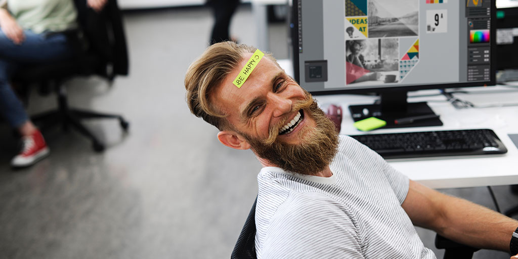 A smiling, bearded man with a note on green paper reading ‘Be happy’ stuck to his forehead.