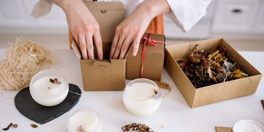 A woman packs brown cardboard boxes using natural materials like straw and leaves. A number of white candles in round containers are on the table.