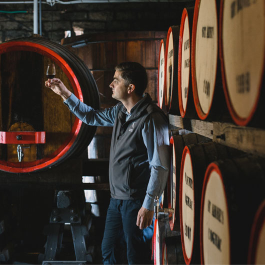A man looking at a glass of wine. He is surrounded by wooden barrels of wine.