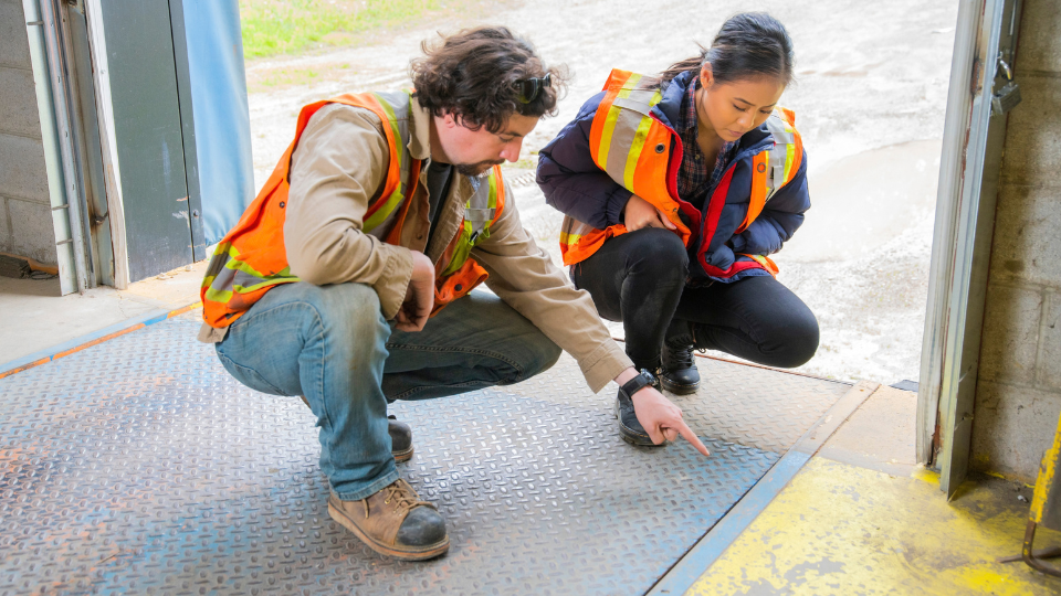 Two workers in high-visibility vests inspecting a warehouse floor surface for a potential safety hazard.