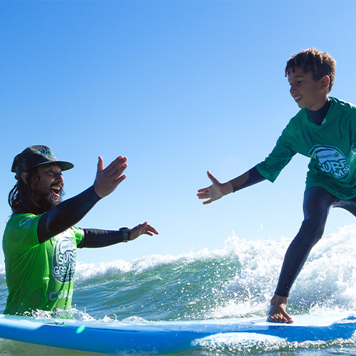 A man standing in the ocean and a young boy standing on a surfboard. Both of them are about to give each other a high five.