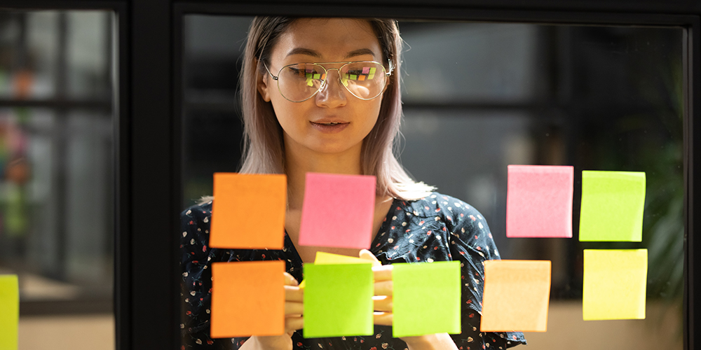 A woman wearing glasses sticks different coloured post it notes on a glass meeting room wall.