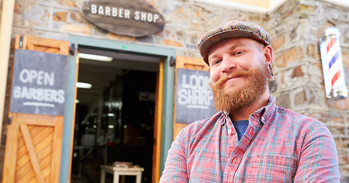 A bearded man wearing a flannel shirt and a driver’s cap stands in front of a barber shop.