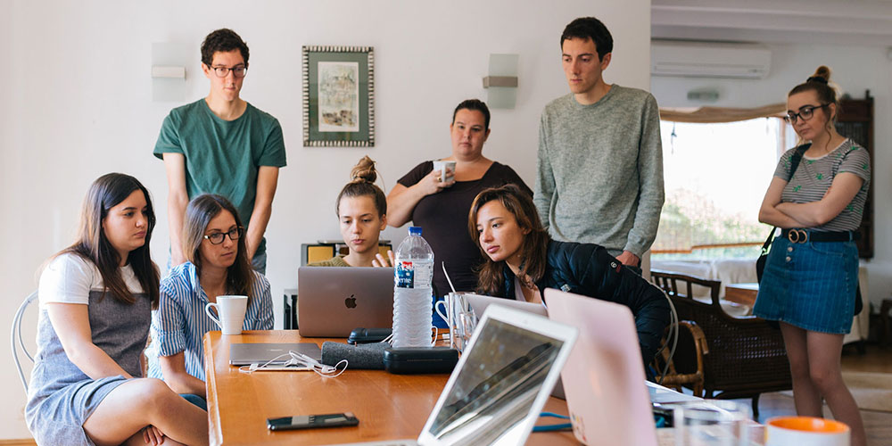A group of six women and two men gather around a laptop for a brainstorming session