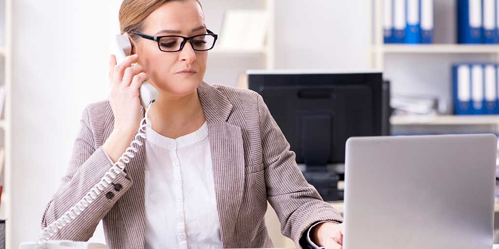 A stern looking woman wearing glasses makes a phone call on a landline telephone with a curly cord.