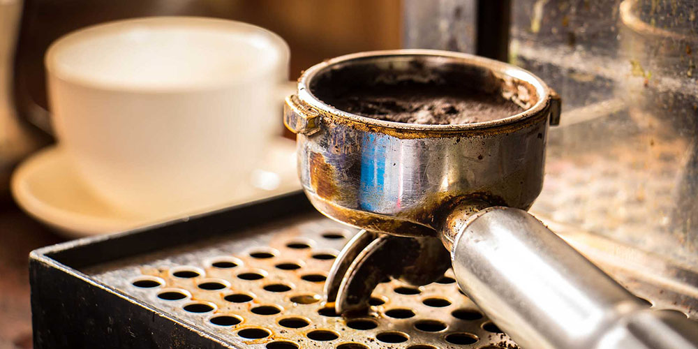 A coffee handle with the the basket full of spent grounds sits on the drop tray of an espresso machine.