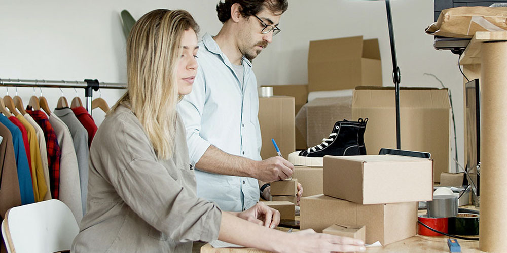 A blonde women sitting in a chair and a dark haired man with facial hair and glasses pack deliveries in brown cardboard boxes.