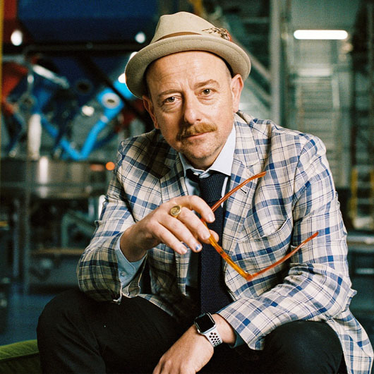 A man wearing a colourful checkered blazer and a fedora, looking at the camera. Behind him is industrial requipment.