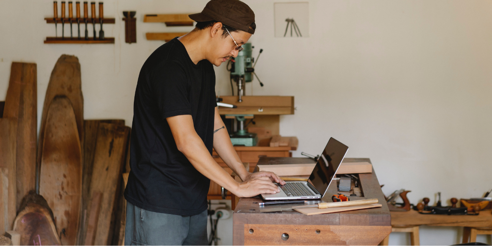 A man standing at a wooden work bench. He is on his laptop. There are work tools and pieces of wood lying around.