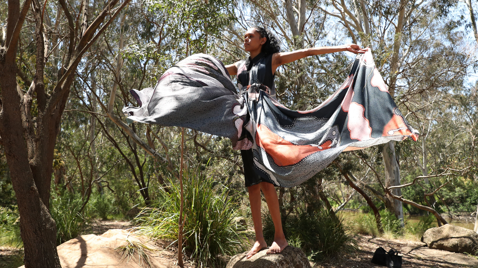 A First Nations woman wearing a dress with Aboriginal colours. She is in the Australian bush.