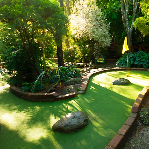 A mini golf course with trees and shrubbery. The mini golf course snakes around with a yellow flag in the hole.