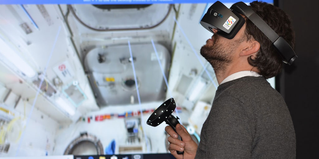 A man wearing a VR headset and holding a controller navigates a virtual version of a space module which can be seen on the screen behind him.