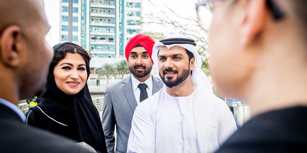 Five people meet outdoors. Of the three people facing the camera, one is a woman wearing a hijab, one a bearded man wearing a suit and a red turban and the last a bearded man wearing white thawb.
