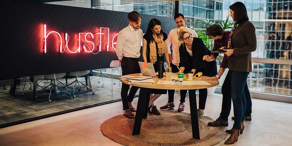 A group of men and women stand around a small, round table. A red neon sign reading ‘hustle’ can be seen on the wall behind them.