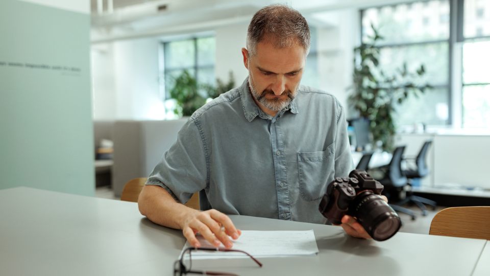 A photographer looking at his camera while sitting at a desk.