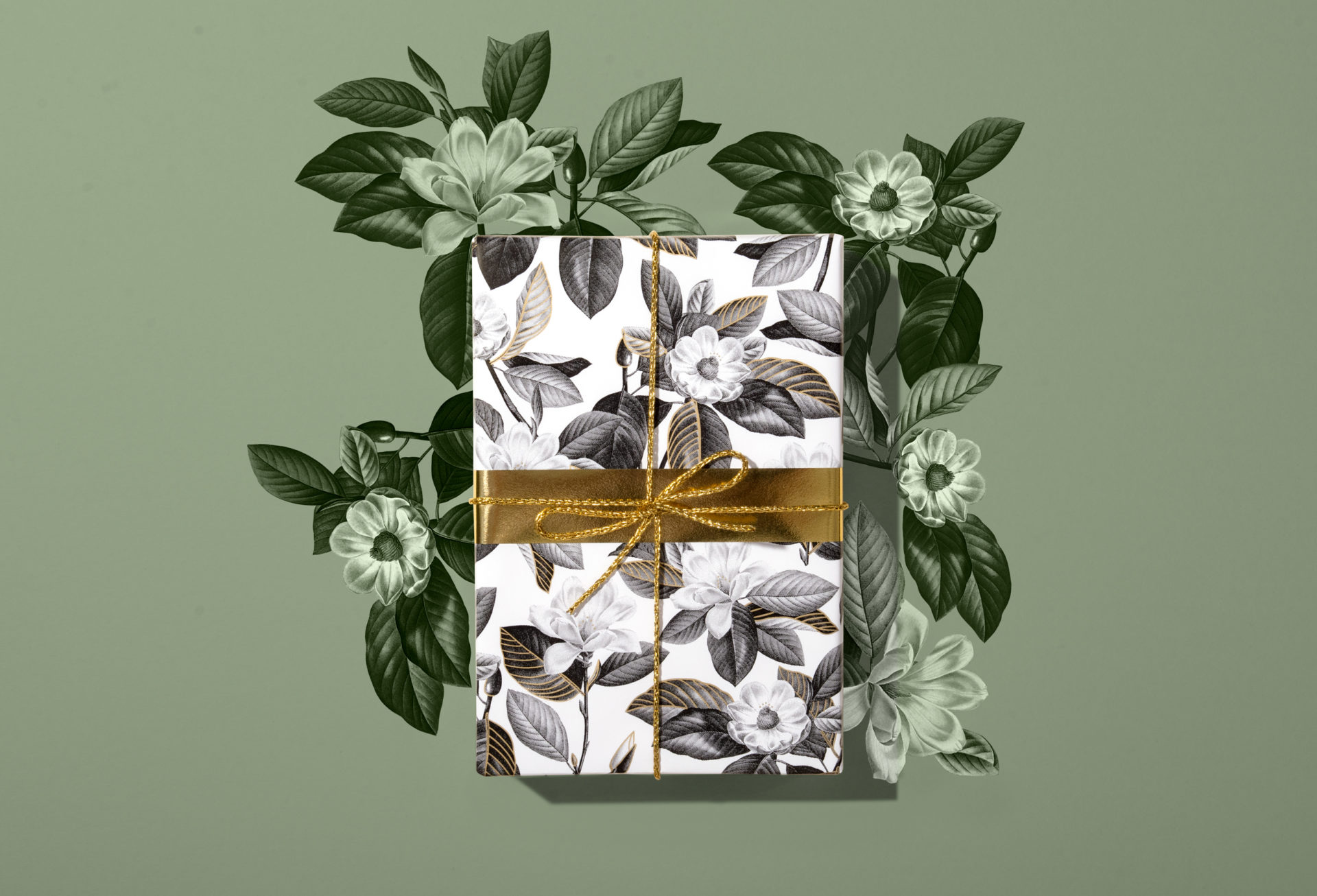 A gift wrapped in black and white wrapping paper with drawings of magnolias, tied with a gold ribbon. Green background echoes the design of the gift wrap. Wrapping paper by Victorian eco-wrapping paper company WrapCo.
