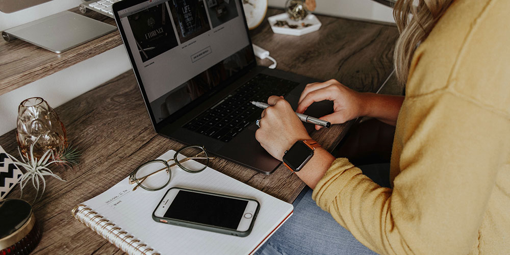 A woman in a hello top works on her laptop which sits on a desk in front of her. She is wearing an Apple Watch and holding a pen. Beside her on a spiral bound notebook are her glasses and an iPhone.