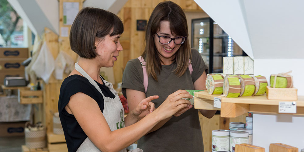 A dark haired shop assistant wearing and gives information about a product to a woman wearing glasses.