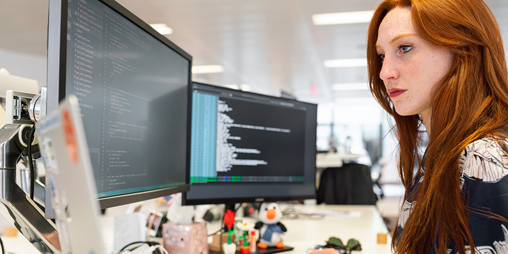 A redheaded woman works on a computer with two screens.