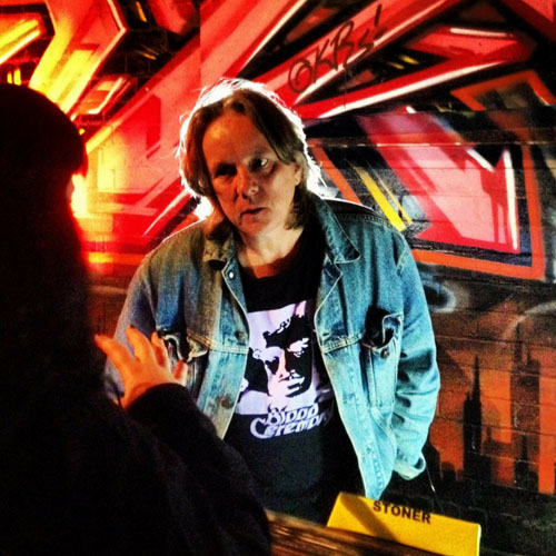 Paul Cook, owner of Heartland Records, stands in front of a striking red patterned graffitied wall and talks to a woman with vinyl records placed between them. He wears a denim jacket and black graphic t-shirt.