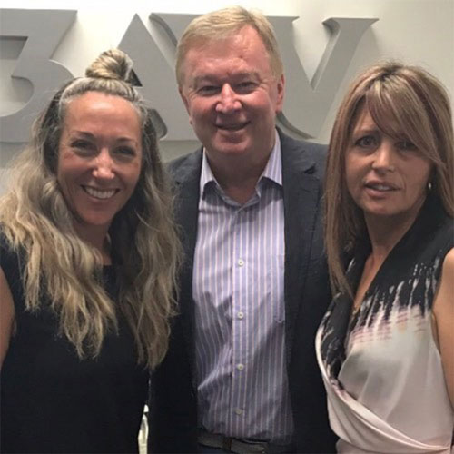 A man and two women, dressed in business casual, smiling as they pose in front of a 3AW wall logo.