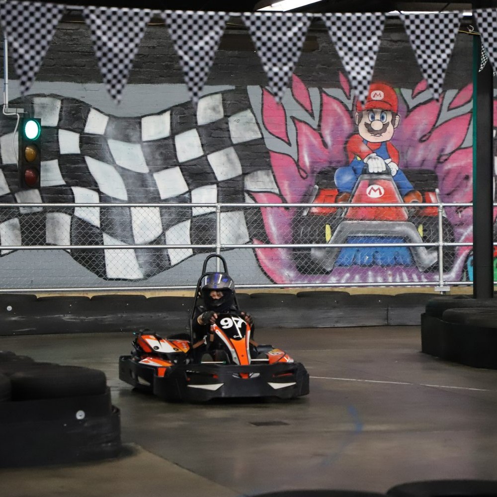 An indoor go-kart circuit. A driver is about to go around a righthand bend. Behind the driver is a cartoon drawing of Mario on the wall.