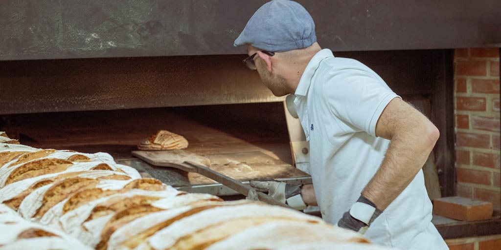 A baker wearing a driving cap, gloves and white top uses a long, wooden paddle to remove bread from an oven.