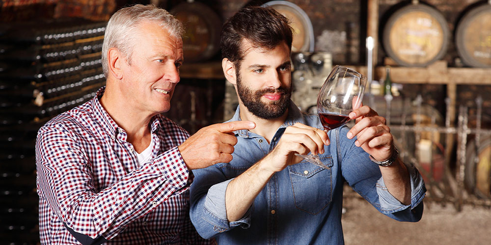 A father and son stand in a wine cellar inspecting red wine in a glass. 