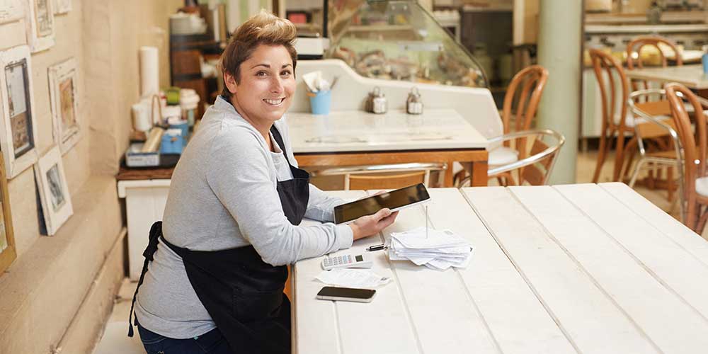 A female shopkeeper wearing an apron. She is holding an iPad. On the counter in front of her is a calculator and a number of receipts on a metal spike. 