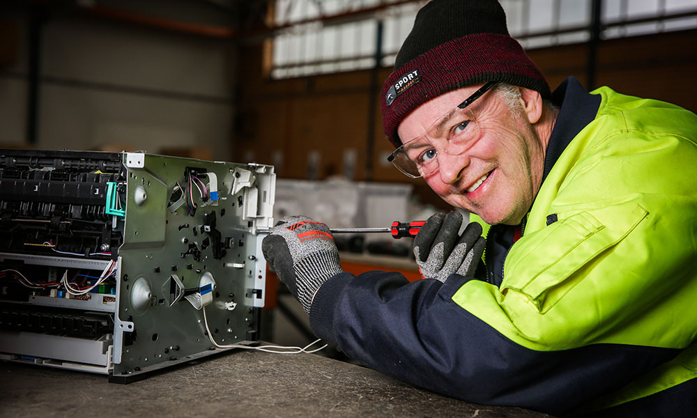 An older man wearing a high viz jacket and beanie opens the casing on a computer using a screwdriver.