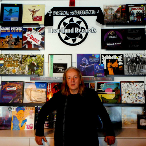 Paul Cook, owner of Heartland Records, stands in front of a wall with the words 'HeartLand Records' painted in white in front of a black graphic sun logo. Numerous colourful vinyl records are displayed on shelves attached to the wall.