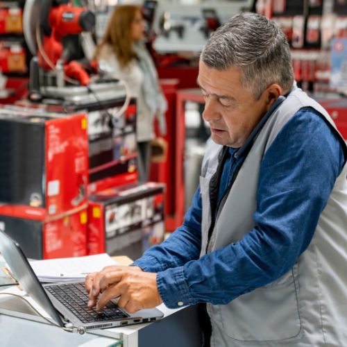 Small business owner using a laptop behind a retail store counter surrounded by tools and equipment.