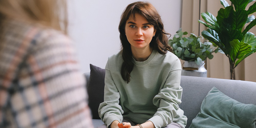 a young woman in dark hair sits on a couch. To the left of frame, we can see the shoulder of her counsellor who wears a checked jacket.