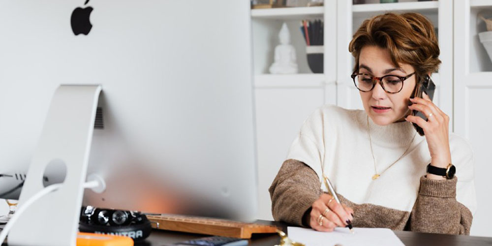 Woman sitting at her desk facing a computer, writing on a notepad whilst on her phone