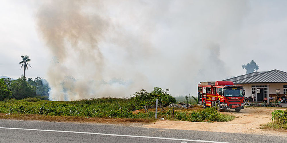 A fire engine is parked in front of a house. In a field to the side of the house, smoke can be seen. 