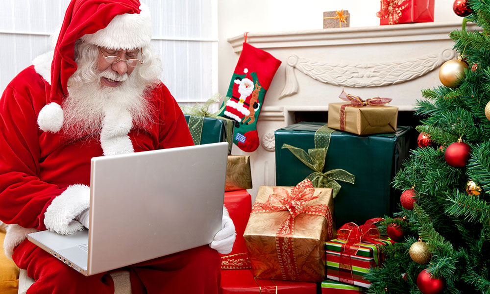 Santa Claus, in his red suit, works on a laptop in a living room filled with presents and a Christmas tree