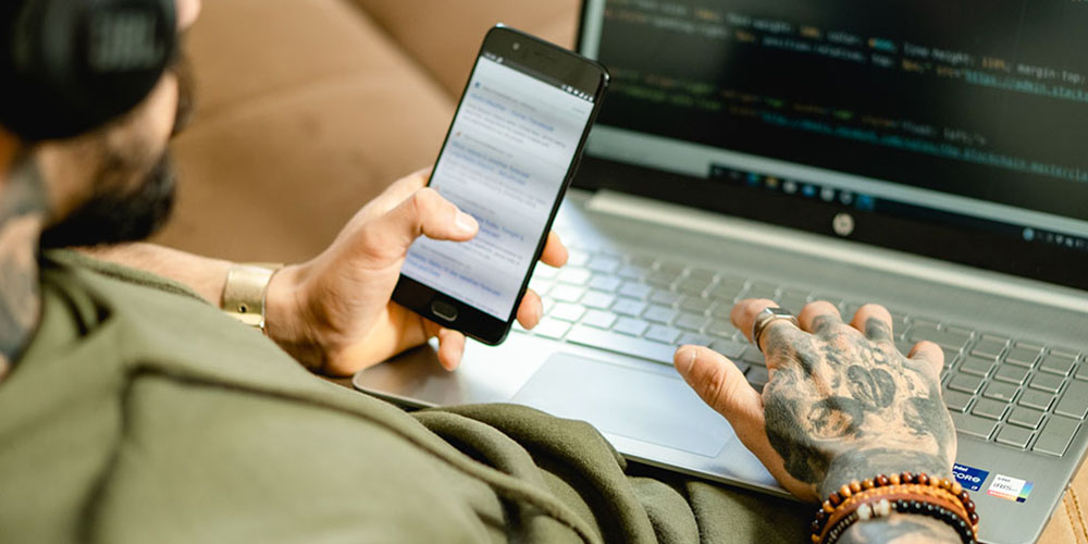 Man with sleeve tattoo scrolling smart phone and laptop wearing headphones