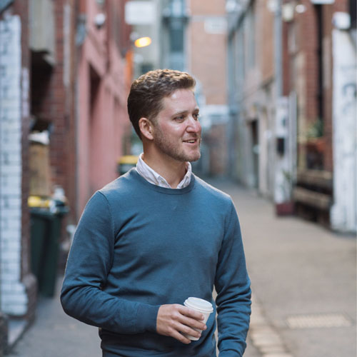 A smiling man with light brown hair wearing a blue sweater over a collared shirt stands in an alleyway holding a takeaway coffee. He looks slightly to his left as though he is listening to someone. 