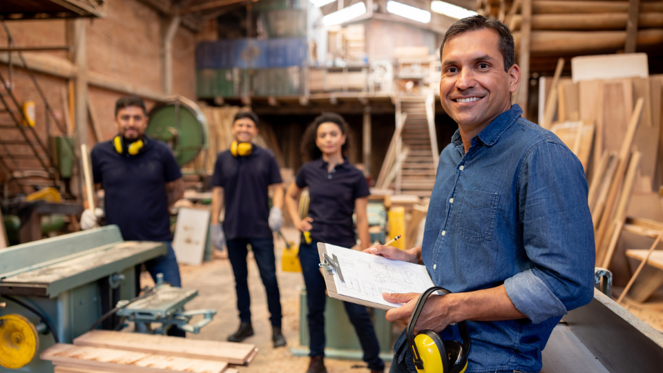 Business manager smiling at a wood factory with his employees at the background