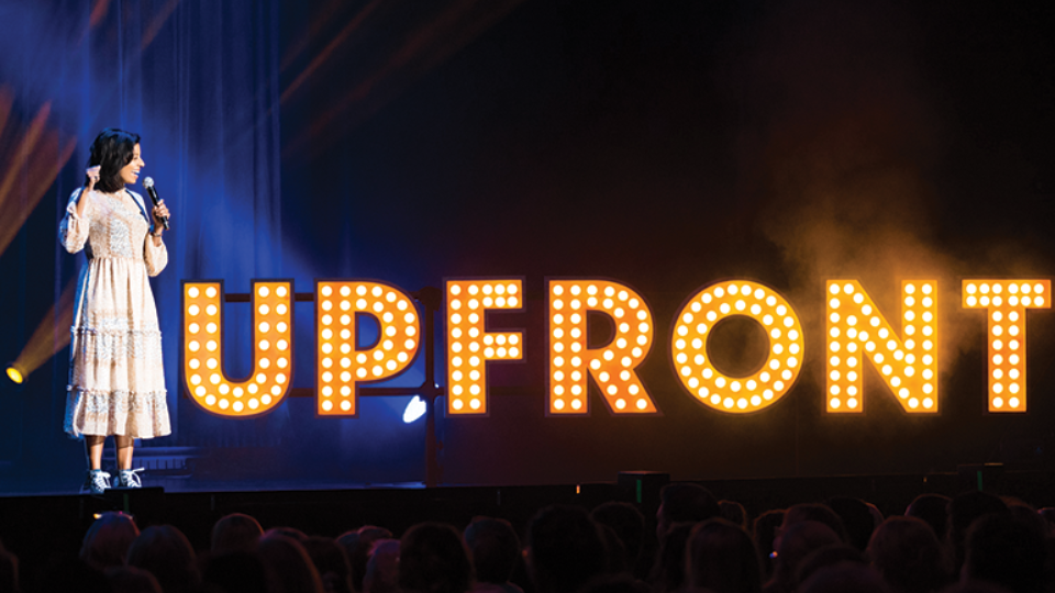 Sashi Perera on stage with a sign reading 'Upfront'.