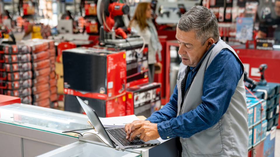 A man in a warehouse on the phone while using a laptop.