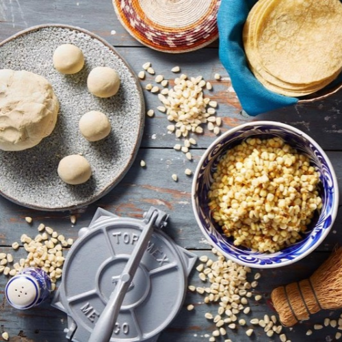 Ingredients and tools used to make tortillas, including corn dough, masa press, corn kernels and stacked tortillas on a table.
