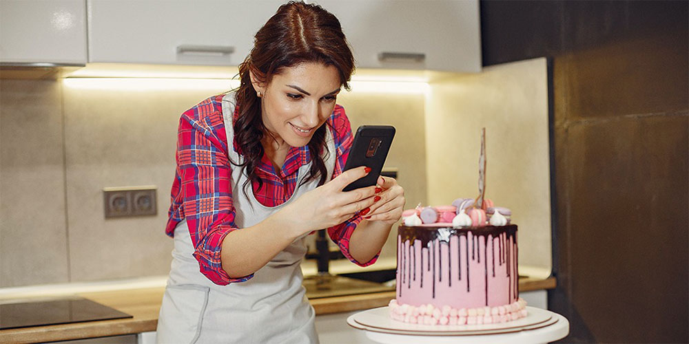 A women wearing an apron stands in her kitchen taking a photograph with her phone of an elaborate looking cake. 