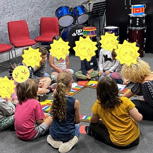 Several kids sitting on the floor playing a musical instrument. Their faces are covered by cartoon suns.